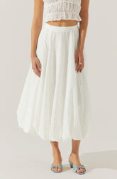 A woman is wearing a white, eyelet, ankle-length skirt with a full, gathered silhouette. She pairs it with a matching white, sleeveless, eyelet top that has a ruffled, cropped design with a smocked waist. She is also wearing pale blue, open-toe sandals with a bow embellishment on the front.